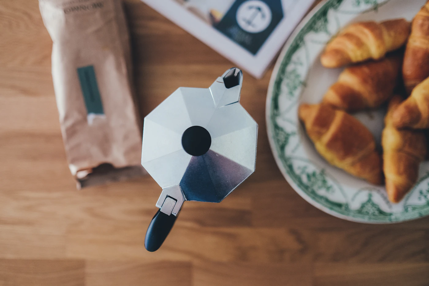 A moka pot and fresh croissants on a wooden breakfast table.
